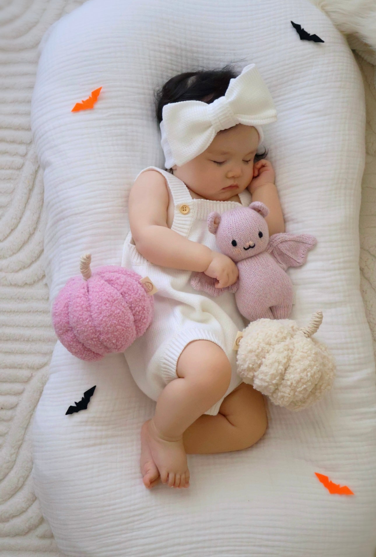 Baby sleeping on a white cushion with stuffed animals, wearing a white outfit and headband.