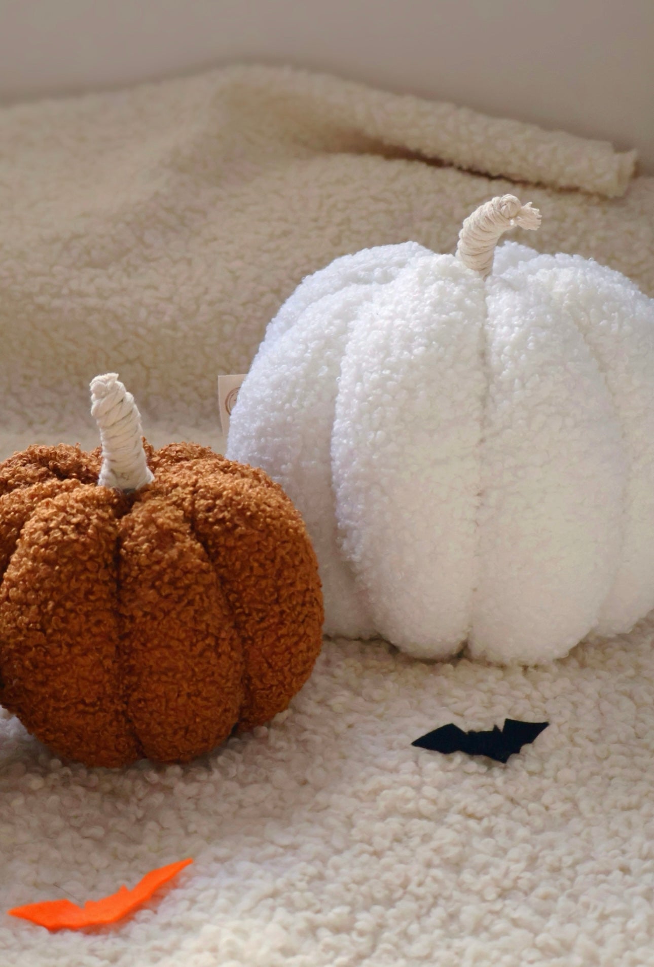 Two decorative pumpkins, one brown and one white, on a textured surface with a small black bat and orange leaf.