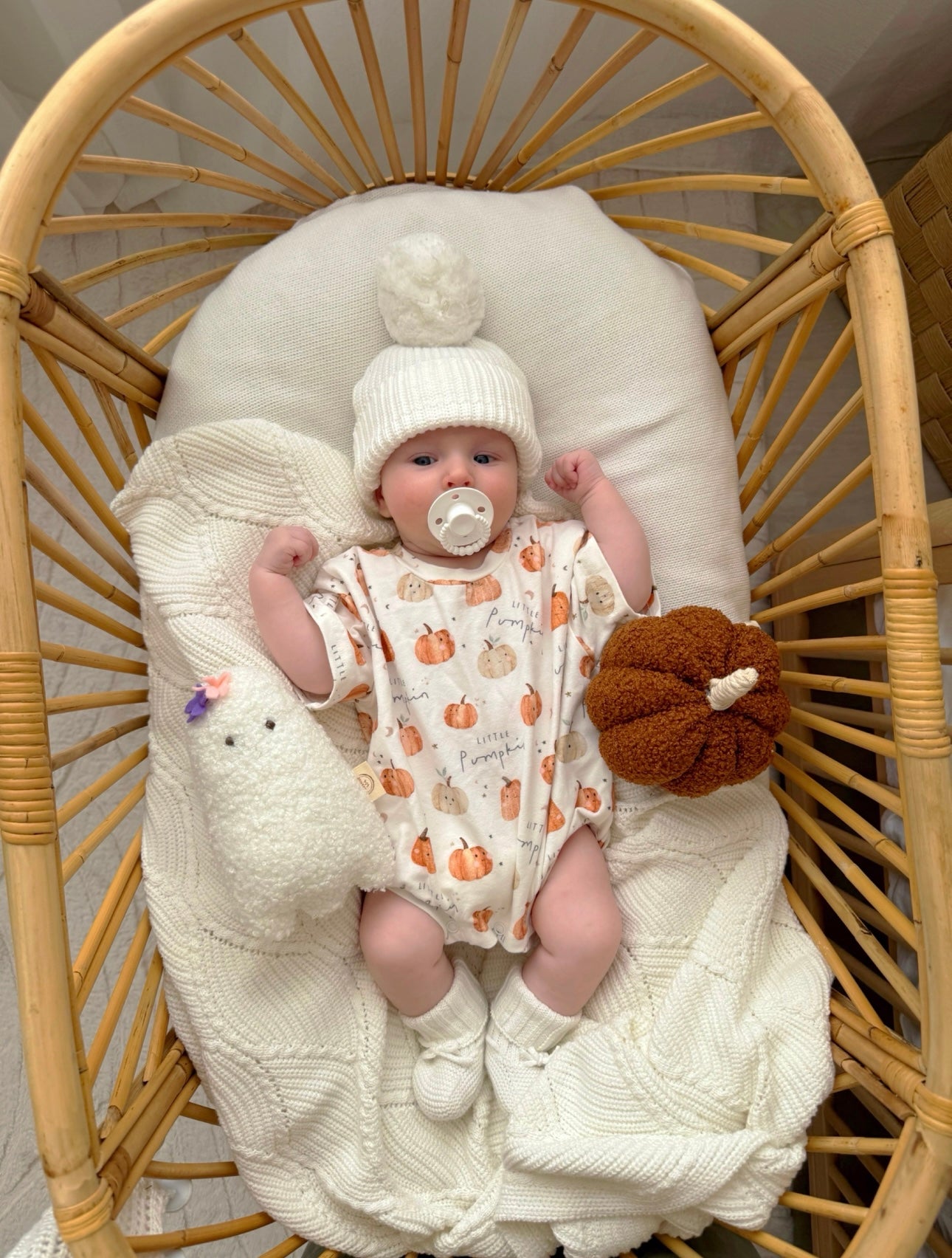 Baby in a crib with a white blanket, pumpkin-patterned outfit, and white hat.