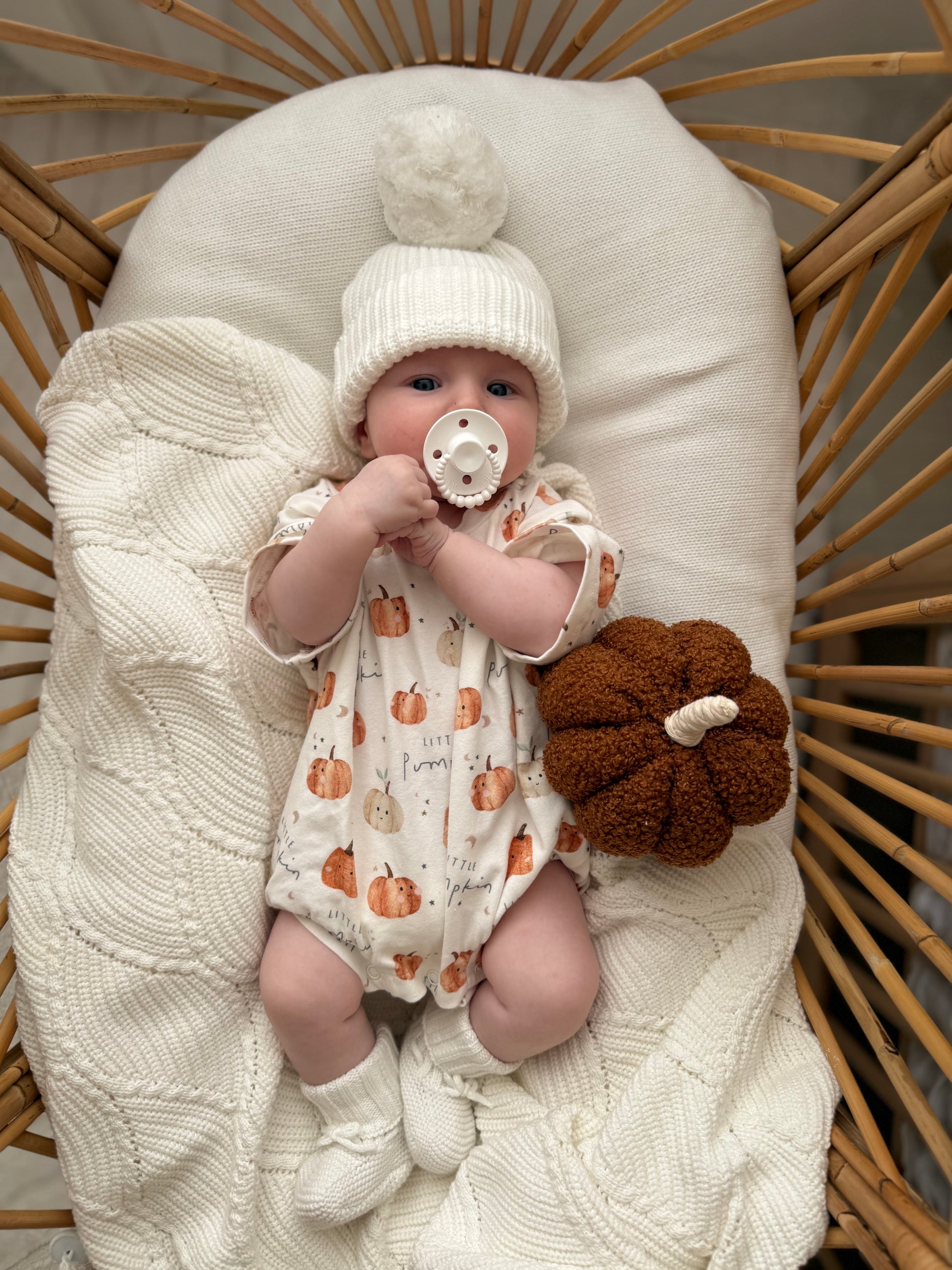 Baby in a crib wearing a white knit hat and pumpkin-patterned outfit, holding a pacifier.