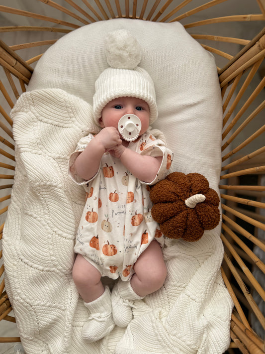 Baby in a crib wearing a white knit hat and pumpkin-patterned outfit, holding a pacifier.